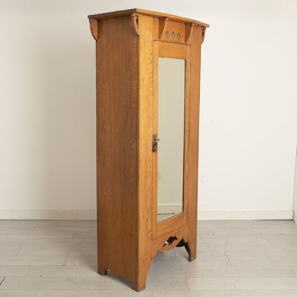 An Arts and Crafts solid oak single door wardrobe featuring a central full-length mirror, carved decorative details beneath an overhanging cornice, and a shaped apron. View II