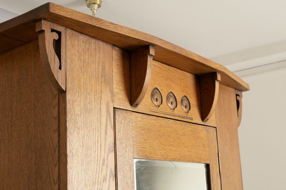An Arts and Crafts solid oak single door wardrobe featuring a central full-length mirror, carved decorative details beneath an overhanging cornice, and a shaped apron. View III