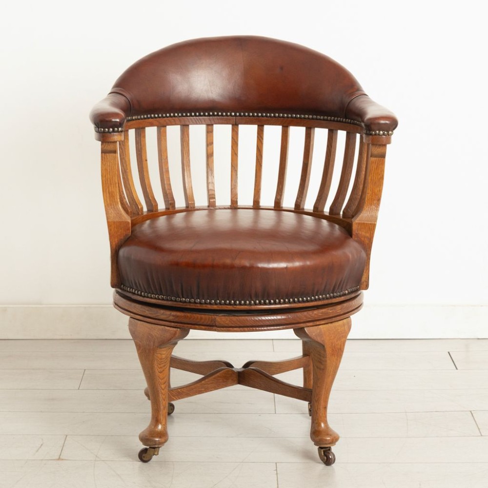 An Edwardian oak swivel library chair featuring a curved spindle backrest, brown leather upholstered seat and top rail with brass studding, resting on cabriole legs with brass castors. View II