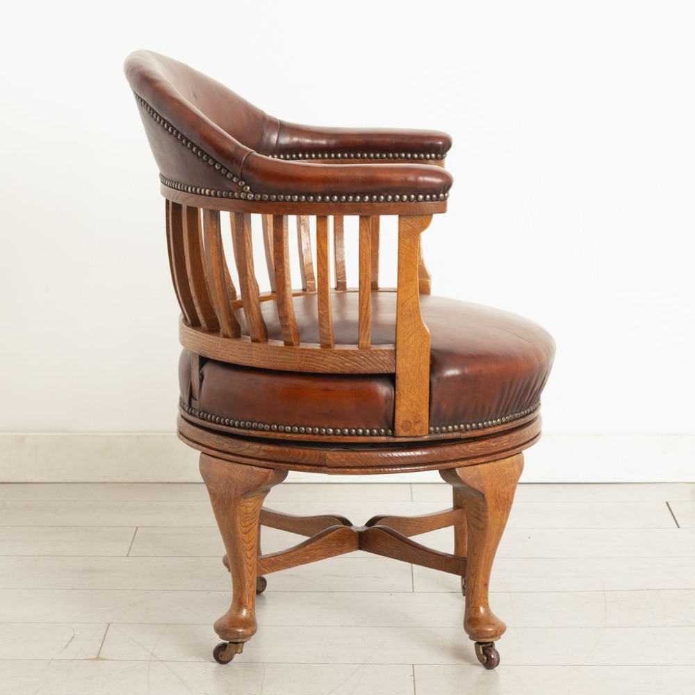 An Edwardian oak swivel library chair featuring a curved spindle backrest, brown leather upholstered seat and top rail with brass studding, resting on cabriole legs with brass castors. View III