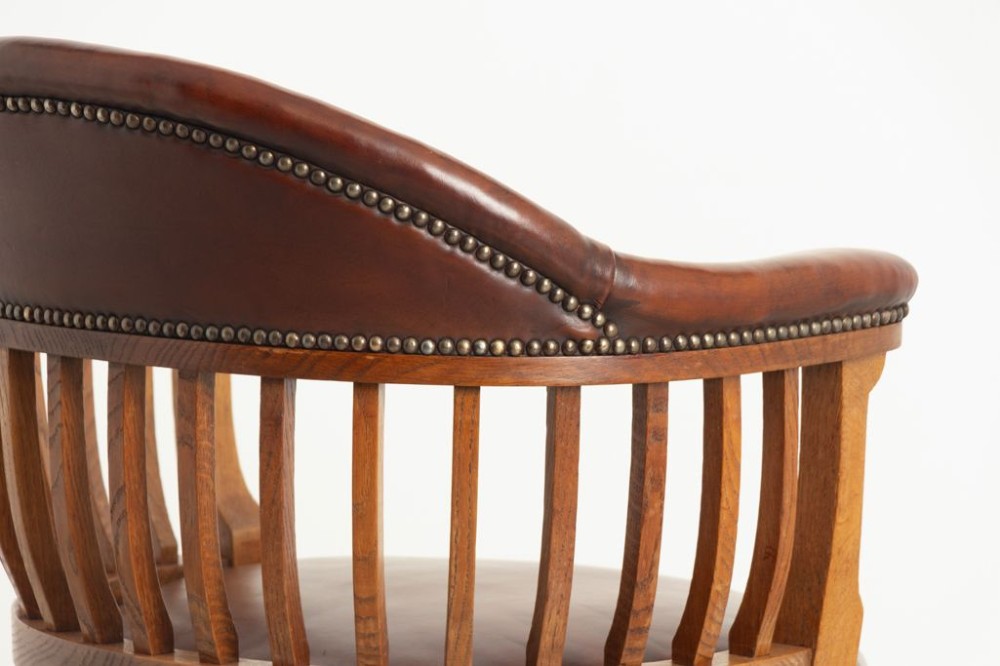 An Edwardian oak swivel library chair featuring a curved spindle backrest, brown leather upholstered seat and top rail with brass studding, resting on cabriole legs with brass castors. View IV