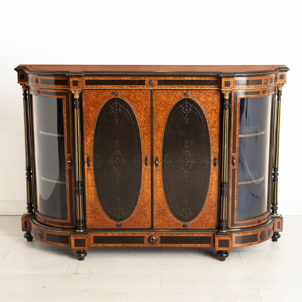 An early Victorian credenza crafted in figure-grained amboyna wood with ebony borders, featuring two central oval-panelled doors flanked by curved glazed side doors and gilt column accents.