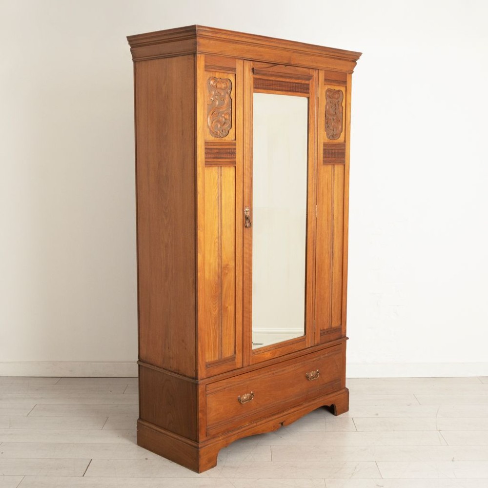 A tall Arts and Crafts walnut wardrobe featuring a central mirrored door flanked by carved timber panels above a single base drawer.