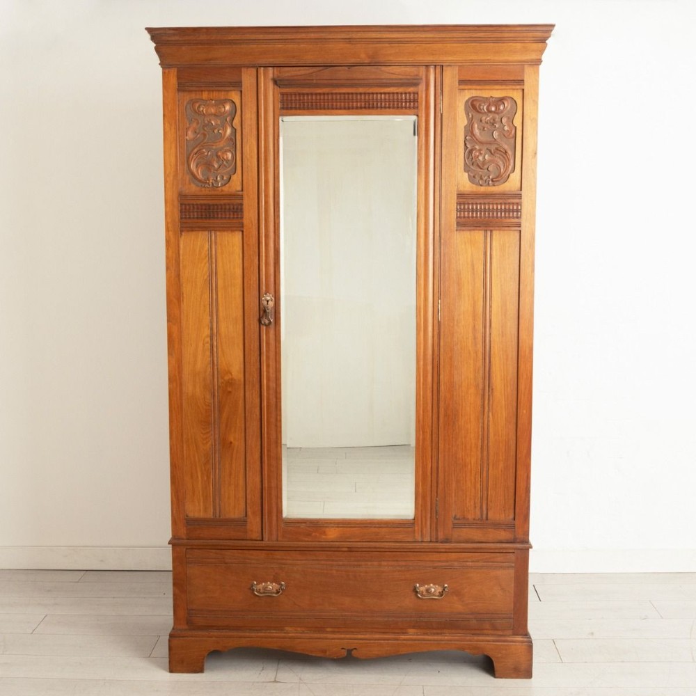 A tall Arts and Crafts walnut wardrobe featuring a central mirrored door flanked by carved timber panels above a single base drawer. View II