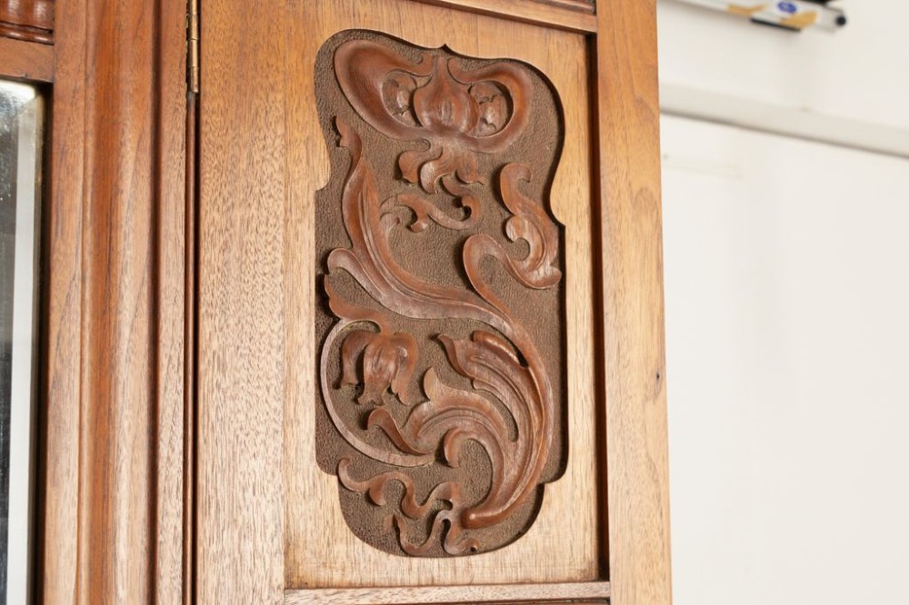 A tall Arts and Crafts walnut wardrobe featuring a central mirrored door flanked by carved timber panels above a single base drawer. View III