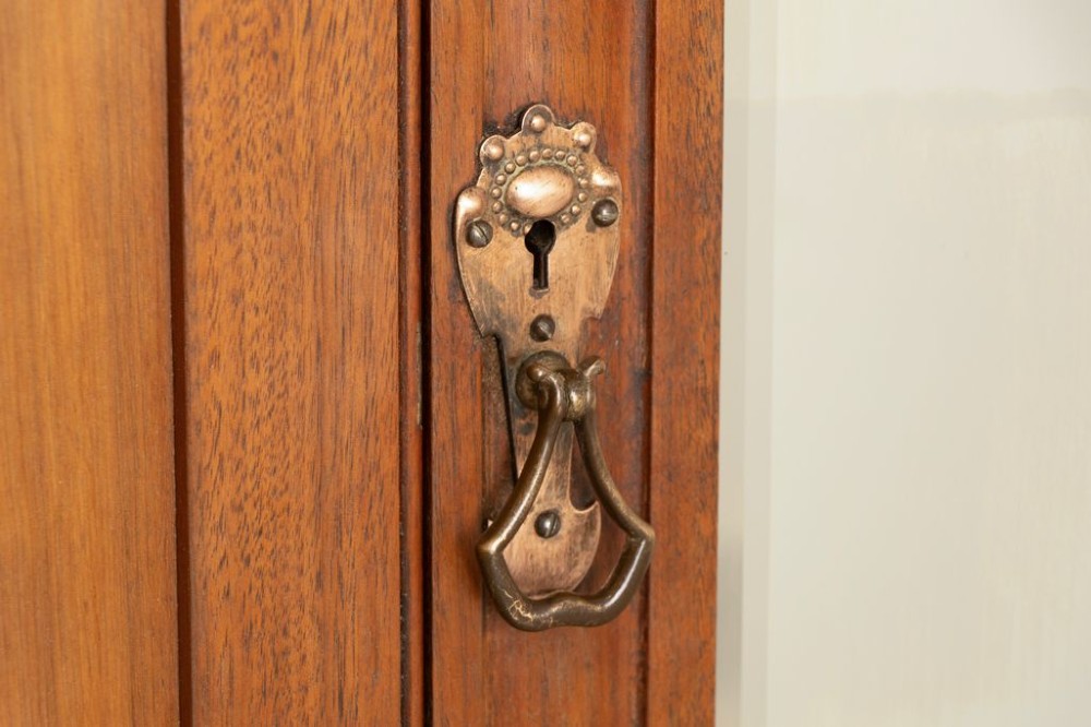 A tall Arts and Crafts walnut wardrobe featuring a central mirrored door flanked by carved timber panels above a single base drawer. View V