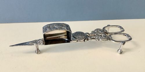 fig 2. An antique silver-plated scissor-style candle snuffer with a central box resting beside a matching oblong tray decorated with engraved floral motifs and scroll borders.