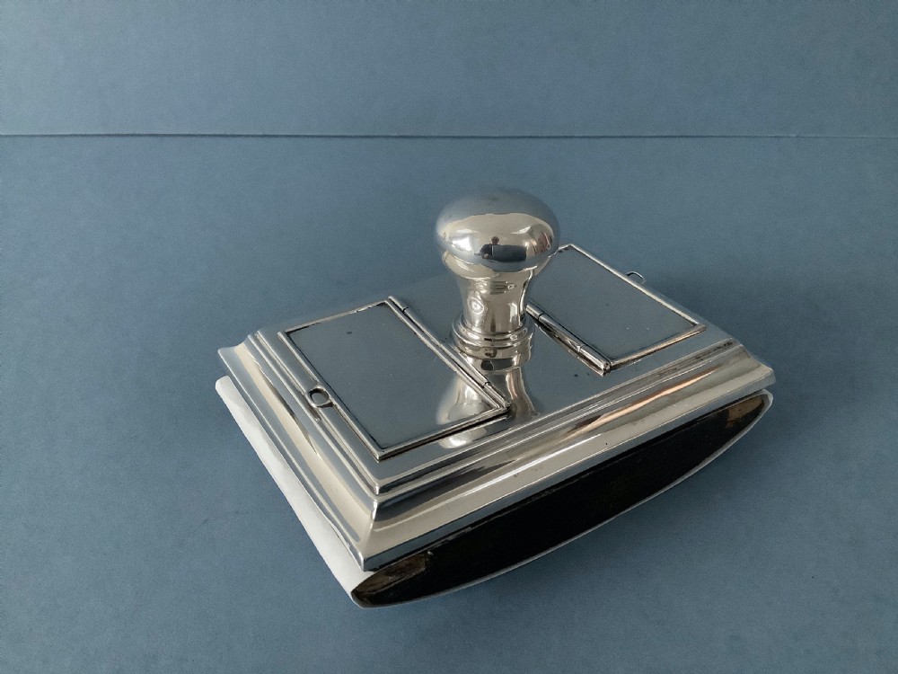 A rectangular silver desk rocker blotter featuring two hinged stamp compartments and a central rounded handle resting on a curved dark base.