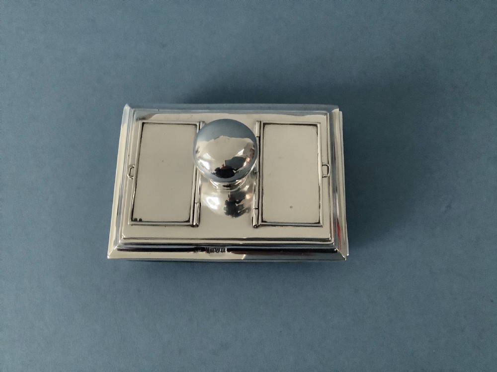 A rectangular silver desk rocker blotter featuring two hinged stamp compartments and a central rounded handle resting on a curved dark base. View II