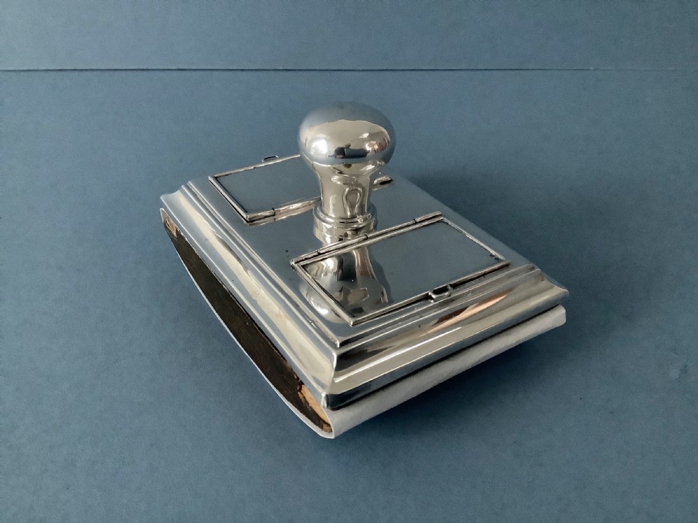 A rectangular silver desk rocker blotter featuring two hinged stamp compartments and a central rounded handle resting on a curved dark base. View V