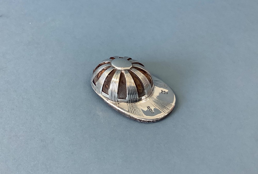 An antique sterling silver pin cushion crafted in the form of a jockey cap with ribbed panels and fabric padding.