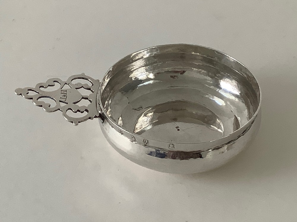 An early 18th-century Britannia standard silver bleeding bowl featuring a shallow round basin and a flat, geometrically pierced handle engraved with initials. View II
