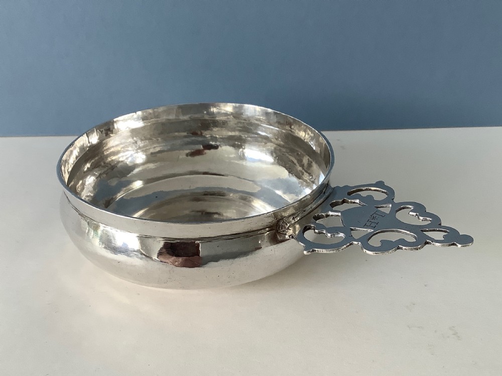 An early 18th-century Britannia standard silver bleeding bowl featuring a shallow round basin and a flat, geometrically pierced handle engraved with initials.