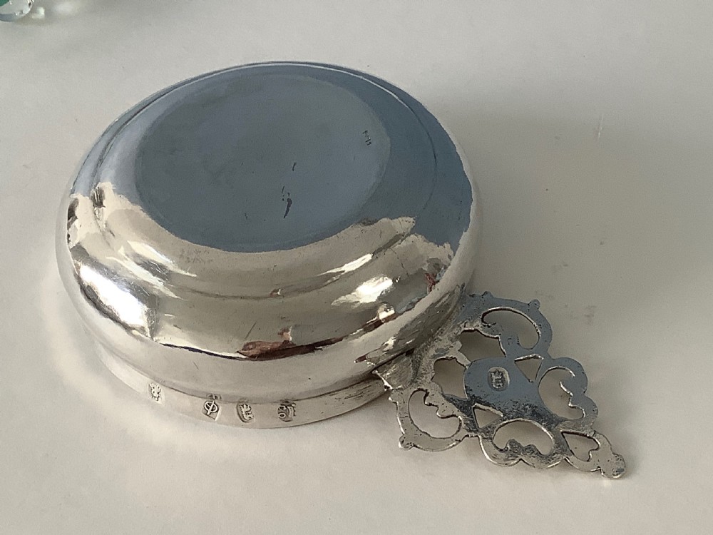 An early 18th-century Britannia standard silver bleeding bowl featuring a shallow round basin and a flat, geometrically pierced handle engraved with initials. View VI