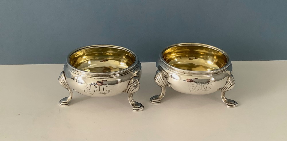A pair of circular Georgian sterling silver open salt cellars featuring gilt interiors, engraved monograms, and tripod legs.