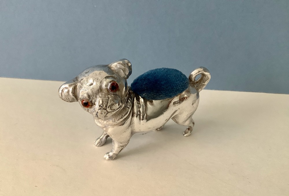 An antique silver pin cushion shaped like a standing pug dog featuring amber glass eyes and a blue velvet cushion set into its back.