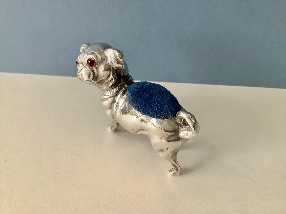 An antique silver pin cushion shaped like a standing pug dog featuring amber glass eyes and a blue velvet cushion set into its back. View II