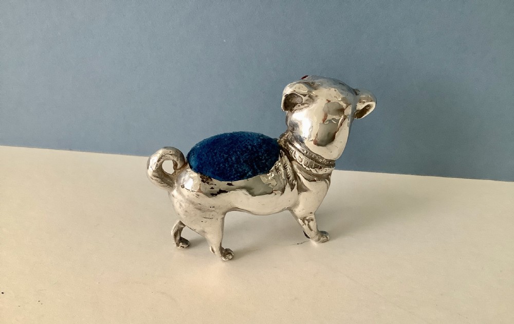 An antique silver pin cushion shaped like a standing pug dog featuring amber glass eyes and a blue velvet cushion set into its back. View III
