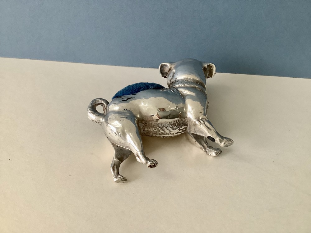 An antique silver pin cushion shaped like a standing pug dog featuring amber glass eyes and a blue velvet cushion set into its back. View V