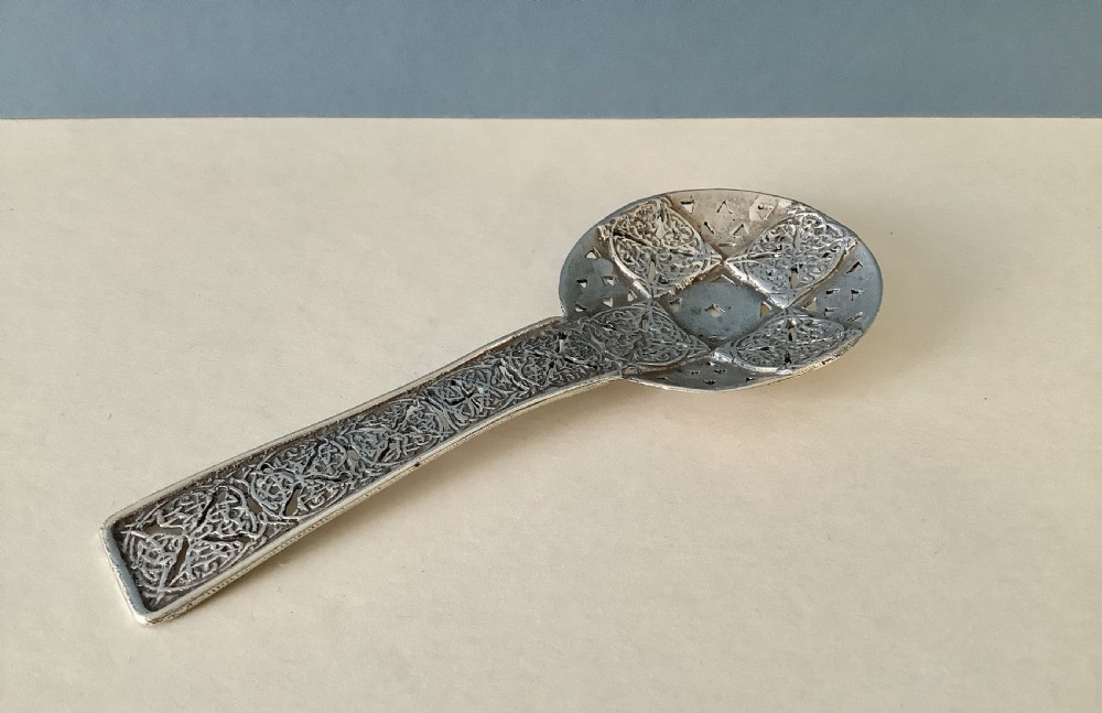 An Arts and Crafts silver caddy spoon featuring a flat circular pierced bowl and a straight handle decorated with intricate knotwork panels. View VI