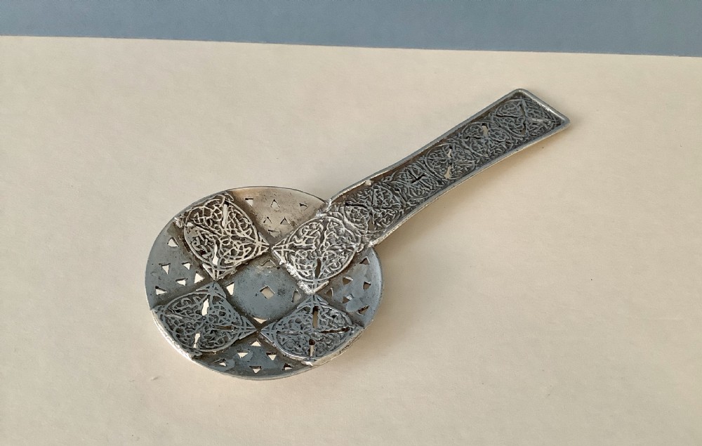 An Arts and Crafts silver caddy spoon featuring a flat circular pierced bowl and a straight handle decorated with intricate knotwork panels.