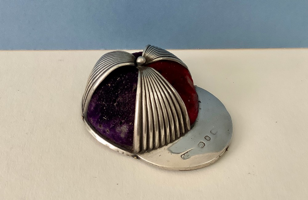An antique silver pin cushion crafted in the form of a jockey cap with ribbed silver details, purple velvet fabric panels, and hallmarks stamped on the front brim.