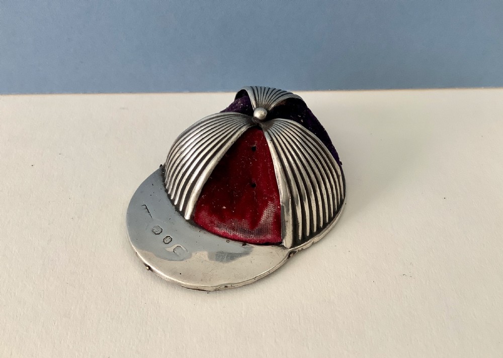 An antique silver pin cushion crafted in the form of a jockey cap with ribbed silver details, purple velvet fabric panels, and hallmarks stamped on the front brim. View II