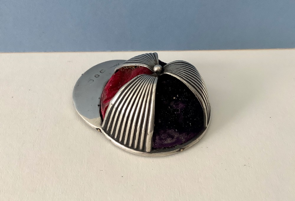 An antique silver pin cushion crafted in the form of a jockey cap with ribbed silver details, purple velvet fabric panels, and hallmarks stamped on the front brim. View III