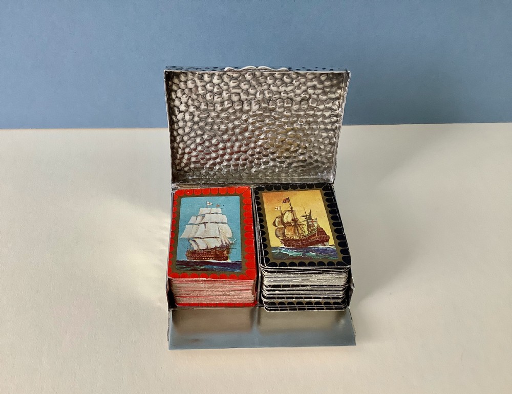 A hammered silver playing card box displayed open with two decks of illustrated playing cards inside. View IX
