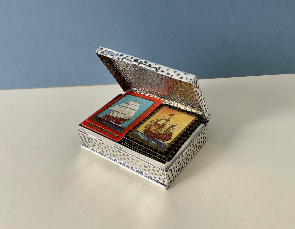 A hammered silver playing card box displayed open with two decks of illustrated playing cards inside.