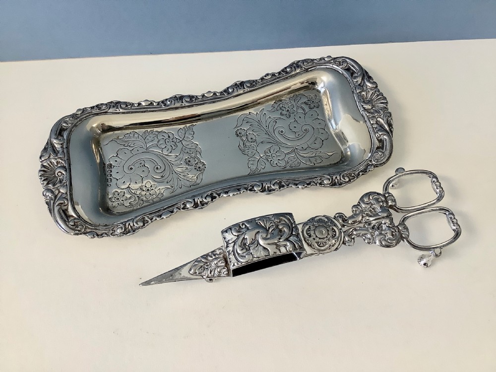 An antique silver-plated scissor-style candle snuffer with a central box resting beside a matching oblong tray decorated with engraved floral motifs and scroll borders.