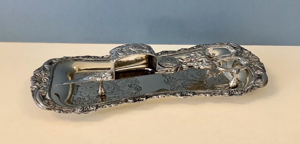 An antique silver-plated scissor-style candle snuffer with a central box resting beside a matching oblong tray decorated with engraved floral motifs and scroll borders. View II