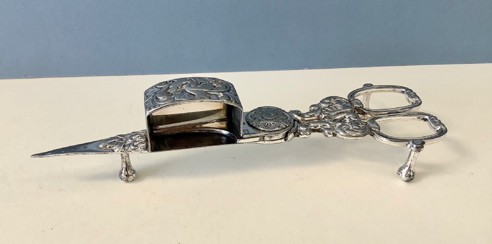 An antique silver-plated scissor-style candle snuffer with a central box resting beside a matching oblong tray decorated with engraved floral motifs and scroll borders. View III