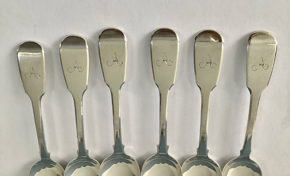 A set of six polished Victorian silver teaspoons featuring engraved monogrammed handles arranged neatly on a plain surface. View V