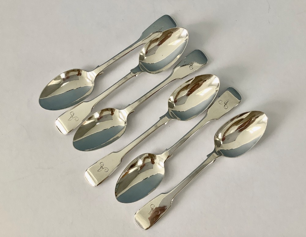 A set of six polished Victorian silver teaspoons featuring engraved monogrammed handles arranged neatly on a plain surface.