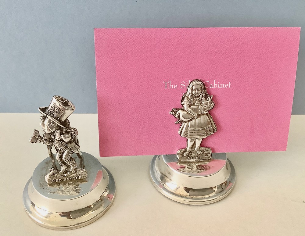 A pair of hallmarked antique silver menu holders depicting Alice and the Mad Hatter standing on round polished silver bases. View IX