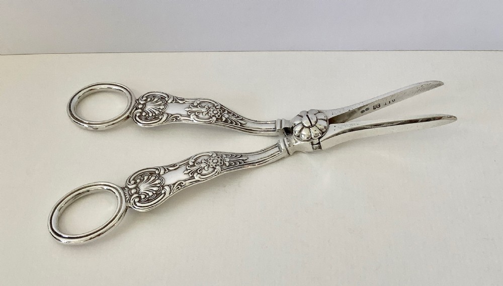 A pair of early Victorian sterling silver grape scissors featuring circular finger handles, chased scroll and shell ornamentation, and a central floral rosette pivot. View V