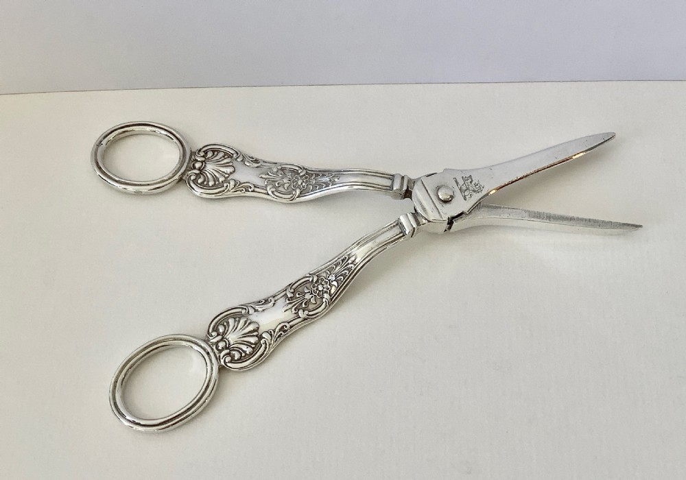 A pair of early Victorian sterling silver grape scissors featuring circular finger handles, chased scroll and shell ornamentation, and a central floral rosette pivot. View VI