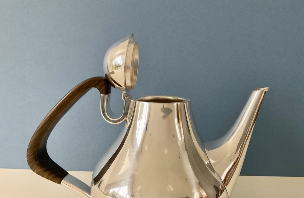 A polished silver Georg Jensen coffee pot featuring a bulbous teardrop body, seamless tapered spout, and a dark carved wooden handle. View V