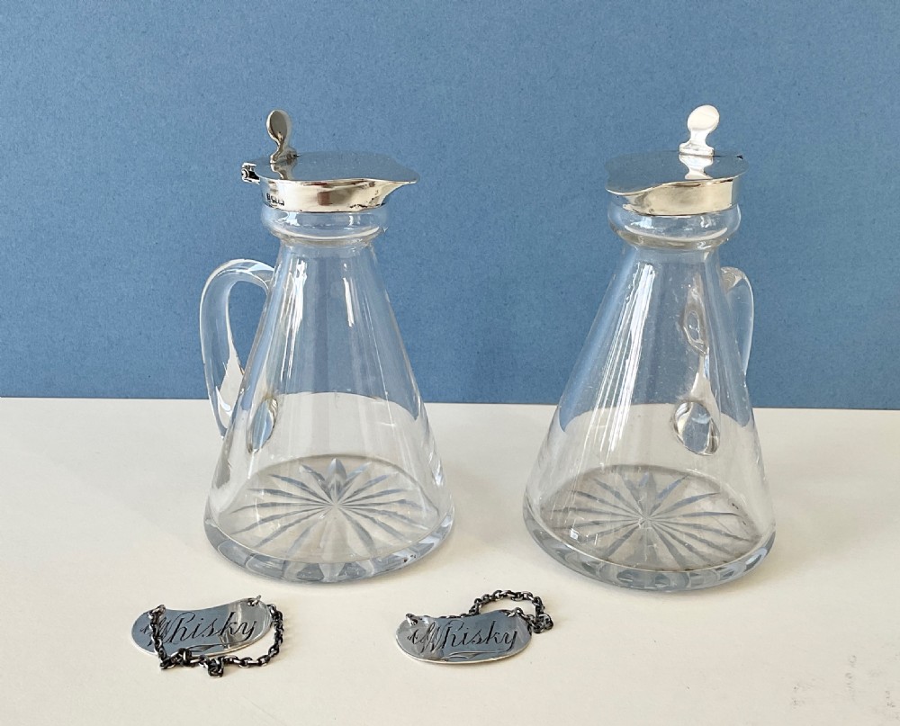 A pair of conical clear glass whisky noggins featuring silver hinged caps, side handles, star-cut bases, and silver Whisky tags.