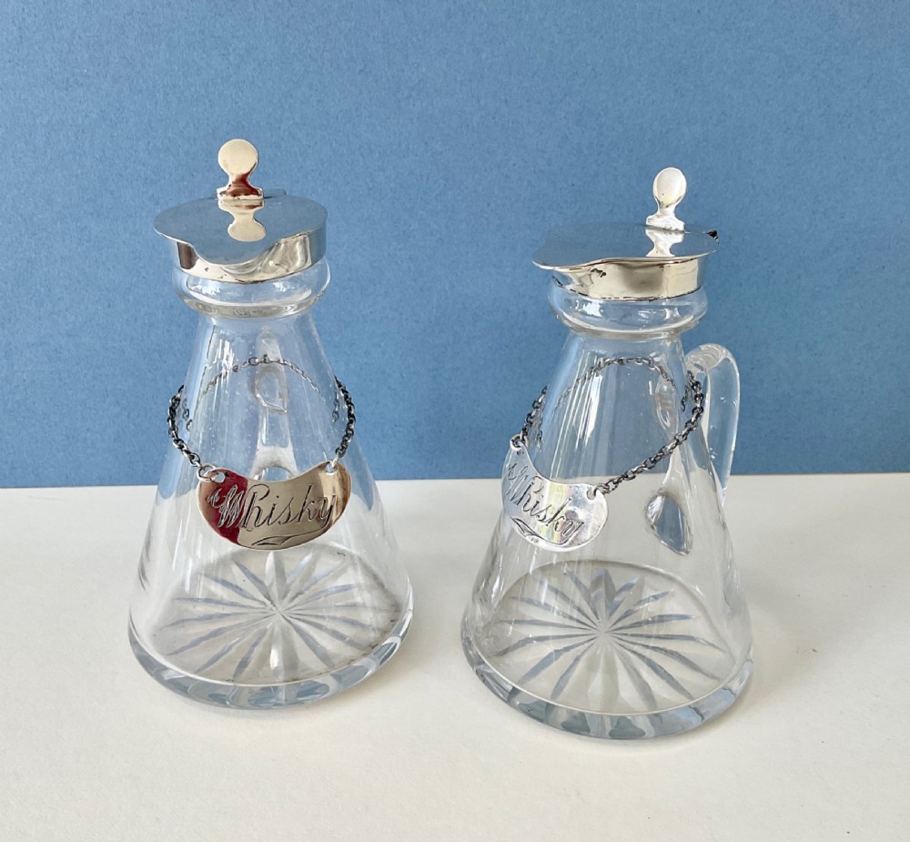 A pair of conical clear glass whisky noggins featuring silver hinged caps, side handles, star-cut bases, and silver Whisky tags. View III