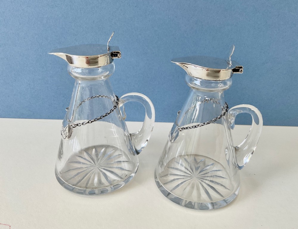 A pair of conical clear glass whisky noggins featuring silver hinged caps, side handles, star-cut bases, and silver Whisky tags. View V
