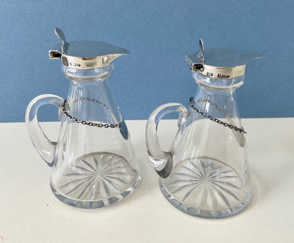 A pair of conical clear glass whisky noggins featuring silver hinged caps, side handles, star-cut bases, and silver Whisky tags. View VIII