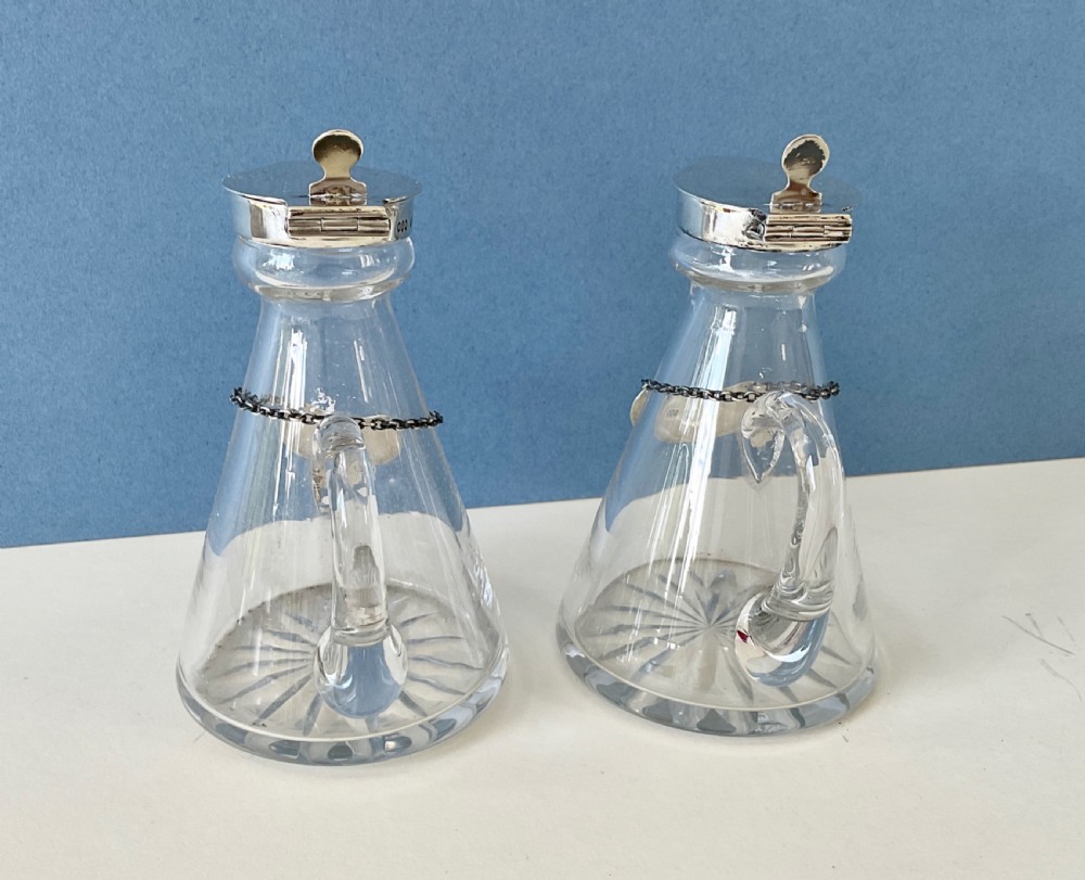 A pair of conical clear glass whisky noggins featuring silver hinged caps, side handles, star-cut bases, and silver Whisky tags. View IX