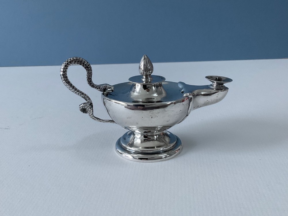 A polished silver table lighter modelled in the shape of an ancient oil lamp with a coiled serpent handle and a pedestal base.