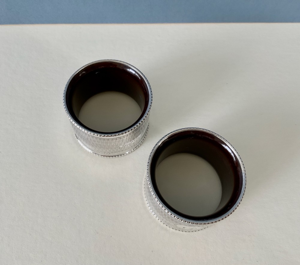 A pair of circular sterling silver napkin rings featuring engine-turned geometric patterns, beaded edges, and dark interior linings. View III