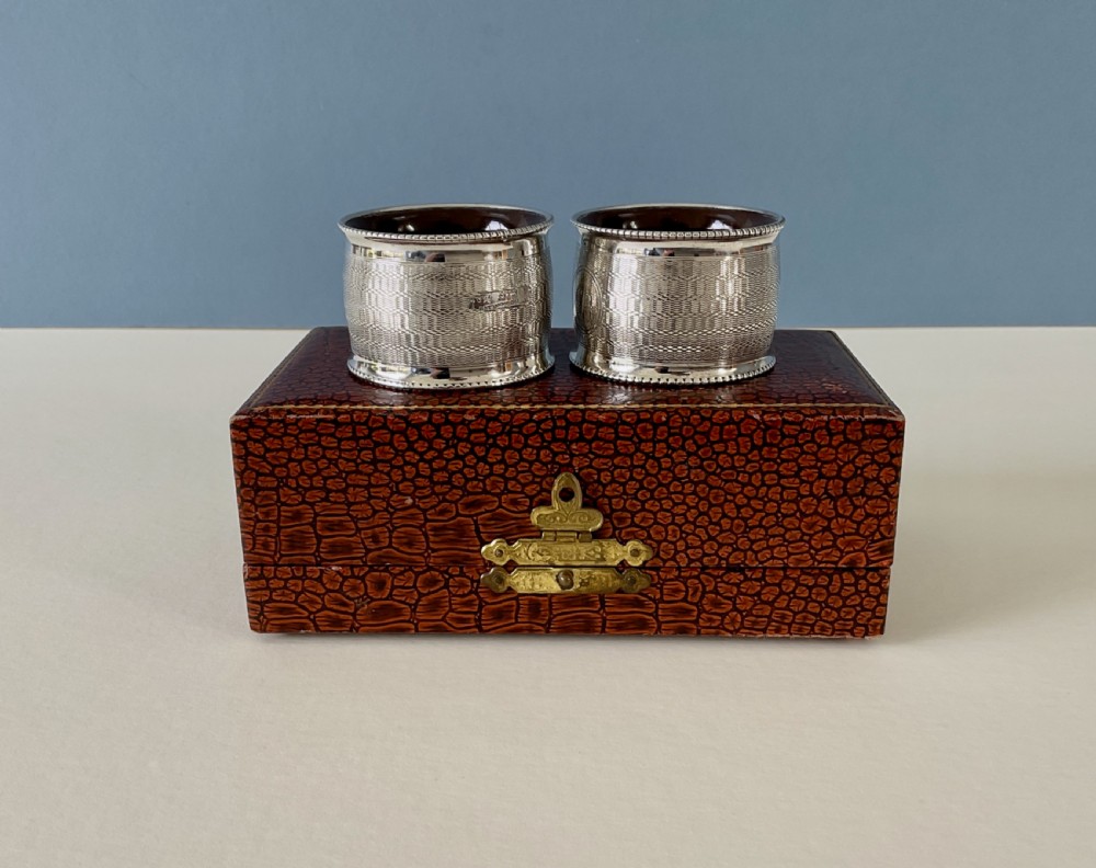 A pair of circular sterling silver napkin rings featuring engine-turned geometric patterns, beaded edges, and dark interior linings. View V