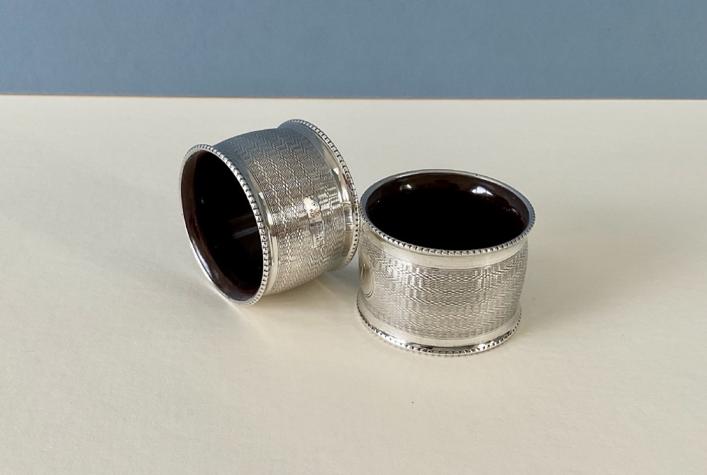 A pair of circular sterling silver napkin rings featuring engine-turned geometric patterns, beaded edges, and dark interior linings. View VIII