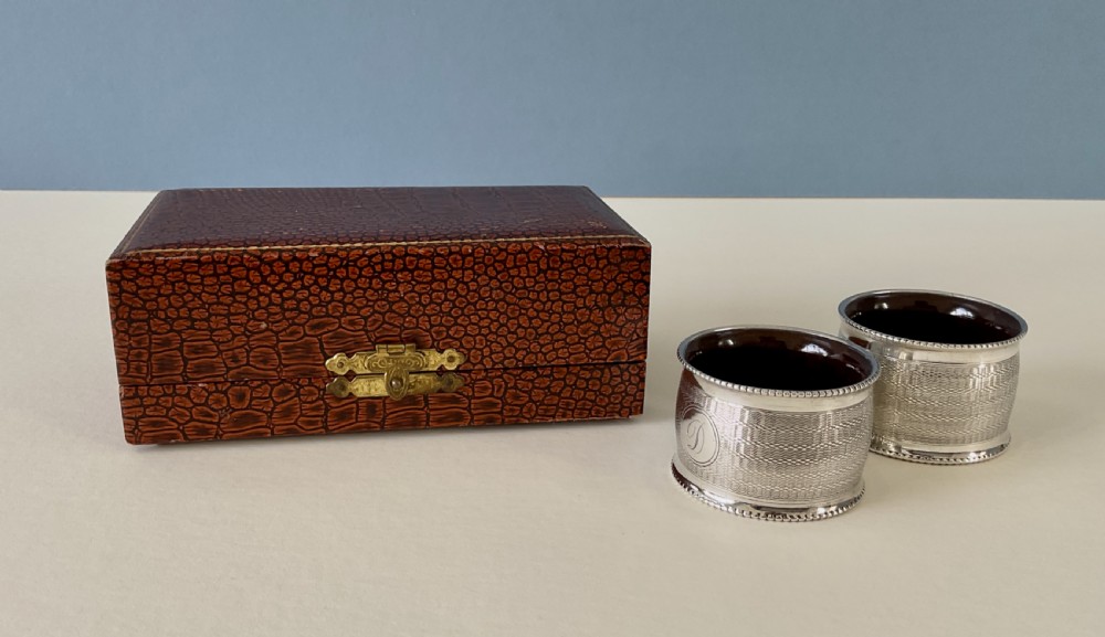 A pair of circular sterling silver napkin rings featuring engine-turned geometric patterns, beaded edges, and dark interior linings. View IX