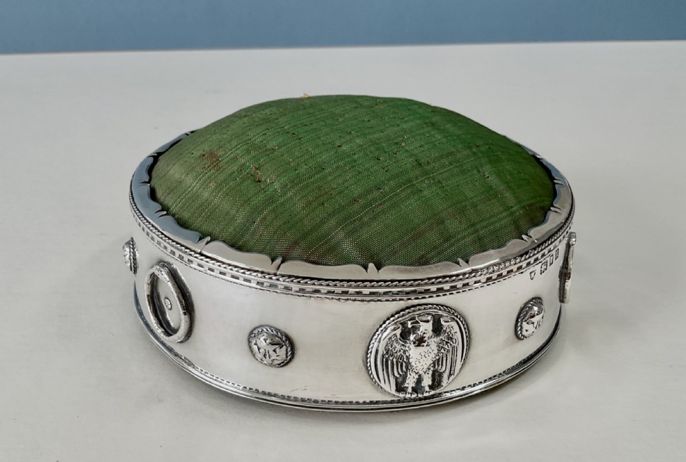 A circular Arts and Crafts silver pin cushion stand with embossed eagle medallions and a raised green woven fabric top.